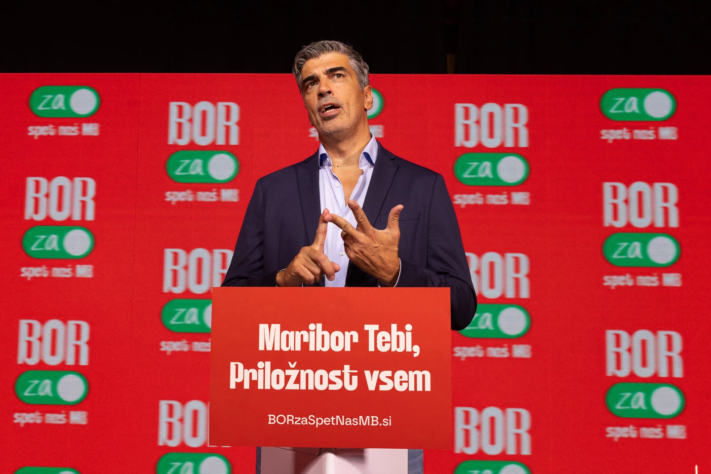 Bor Greiner govori za pultom pred rdečim kampanjskim ozadjem.