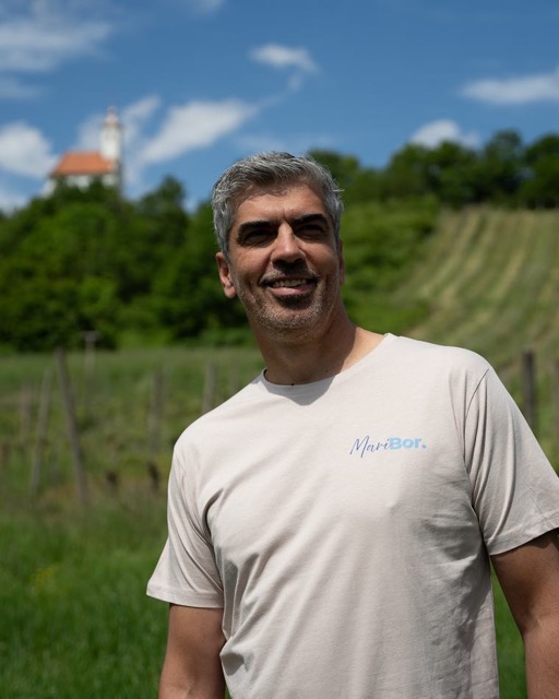 Bor Greiner v majici stoji pred vinogradom in cerkvijo na griču.